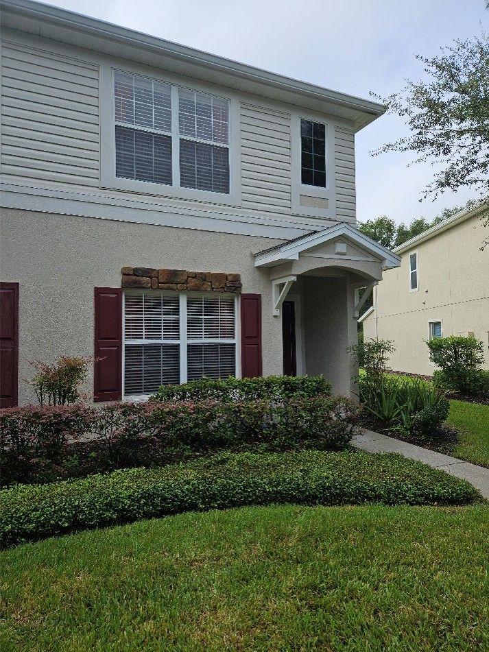 Two-story beige house with white trim, red shutters, and a small covered front porch, bordered by a green lawn.