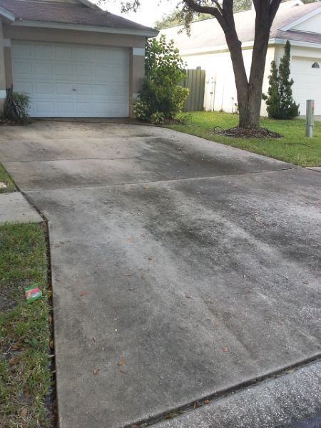 A gray, stained concrete driveway leading to a closed garage door with a lawn and tree nearby.