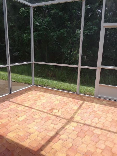 Green screened-in patio/courtyard beside a hedge and wet lawn, with potted plants and a stone path.