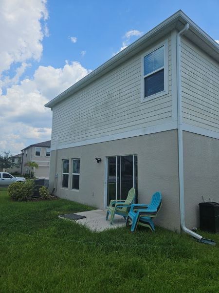 A two-story house with cream-colored siding and stucco, featuring a patio with two plastic chairs on a grassy yard.