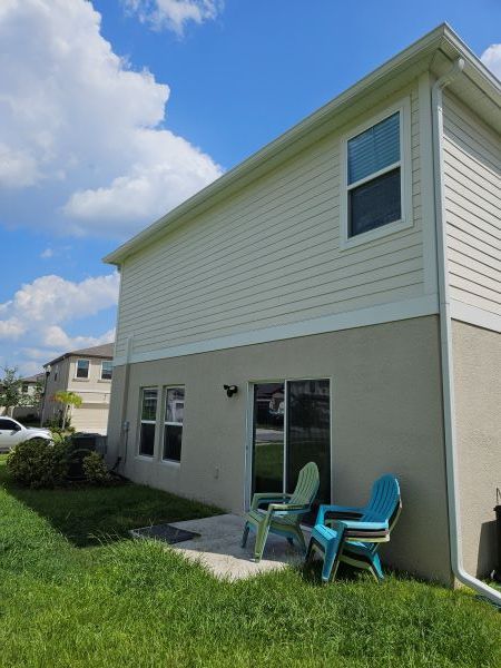 A two-story house with beige stucco on the lower level and light siding above, featuring a small patio with two chairs.