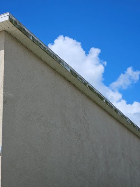 A tan stucco exterior wall under a white roof eave against a vibrant blue sky with fluffy white clouds.