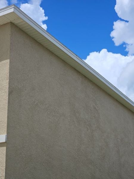 A tan stucco exterior wall under a white roof eave against a vibrant blue sky with fluffy white clouds.