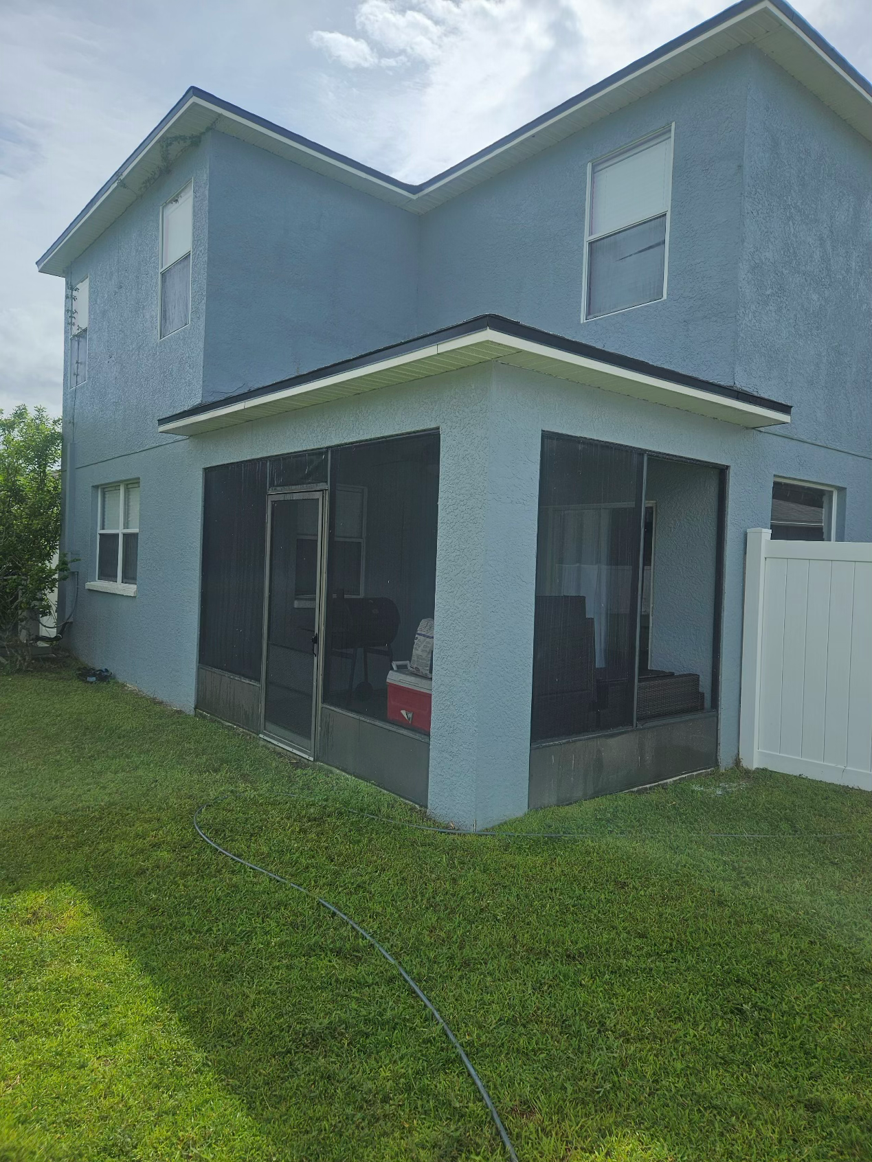 A light blue two-story house with a screened-in porch on the ground floor and a white fence in a green grassy yard.