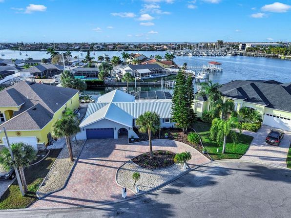 Aerial view of waterfront houses with palm trees and a harbor in the background under a blue sky.