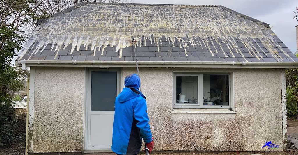Person in blue jacket cleaning a small, dirty building's roof with streaky, white stains on a cloudy day.