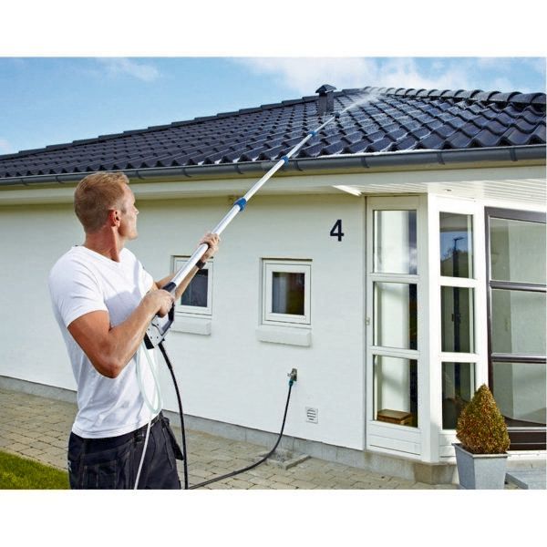 Man pressure washing a roof with an extendable wand, white house, blue sky.
