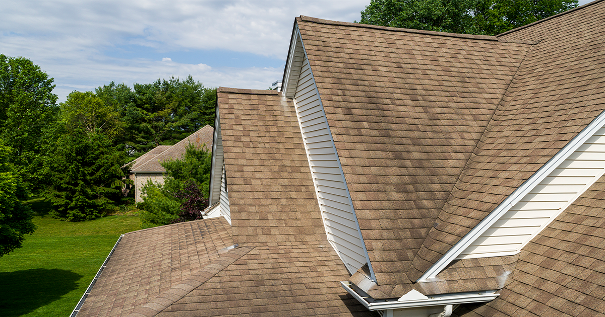 Brown shingled roof with a dark stain, white trim, and a backdrop of green trees and a lawn.