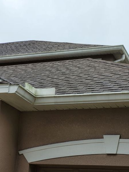 Tan house with brown shingled roof, white gutters and trim under overcast sky.