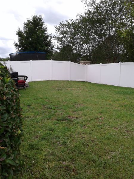 Grassy backyard with a white vinyl fence, trees, and a trampoline in the corner