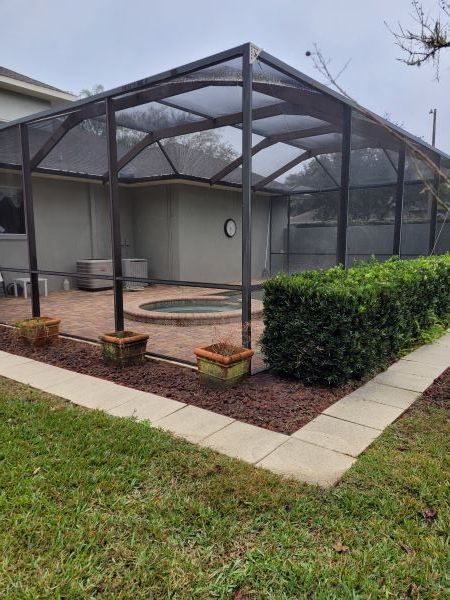 Green screened-in patio/courtyard beside a hedge and wet lawn, with potted plants and a stone path.