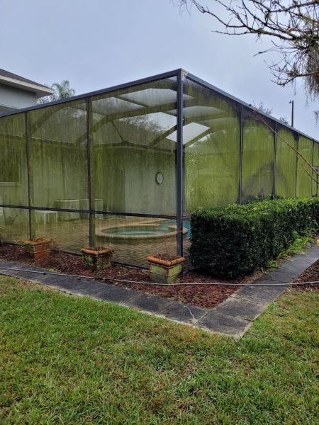 Green screened-in patio/courtyard beside a hedge and wet lawn, with potted plants and a stone path.