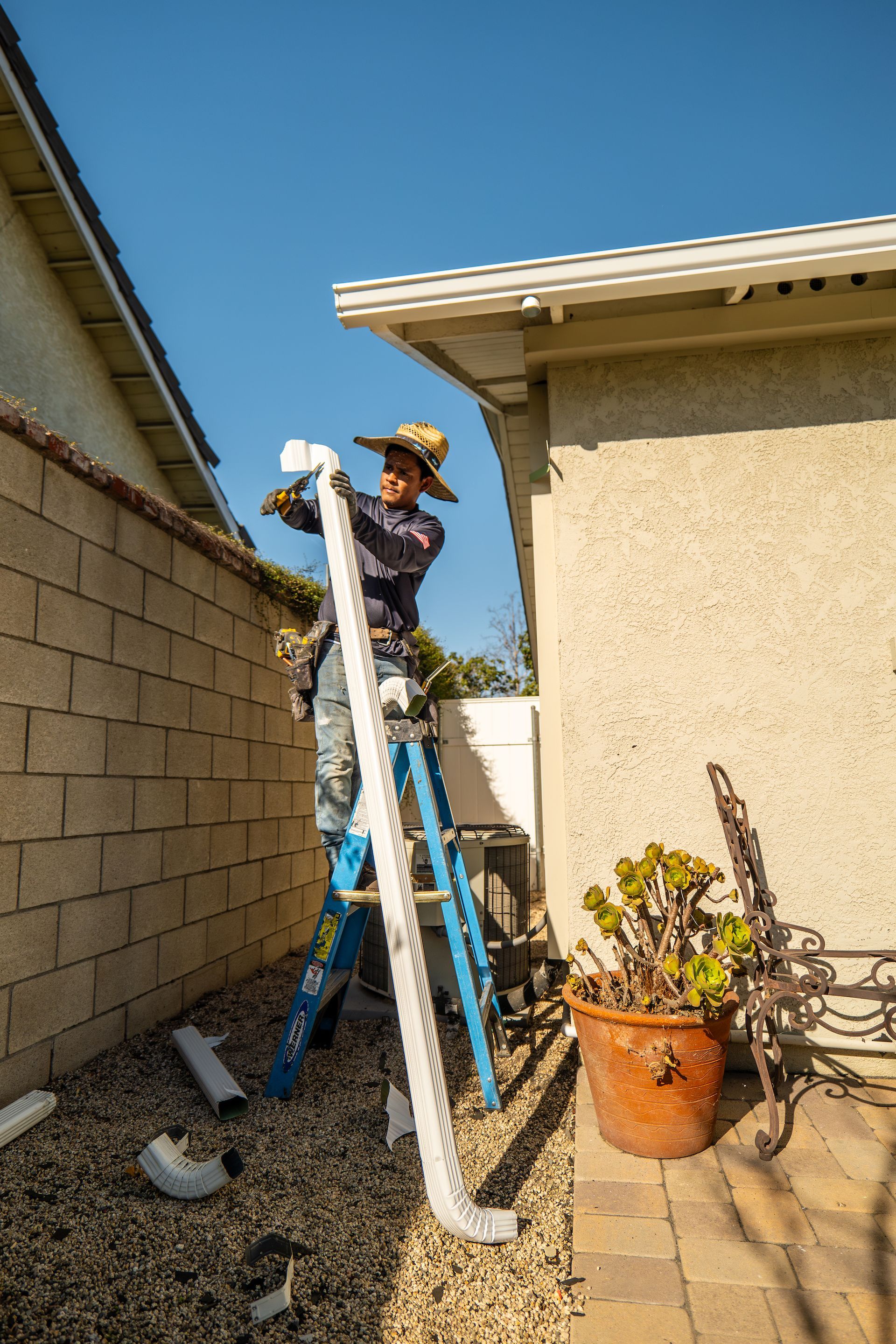 A man is standing on a ladder working on a pipe outside of a house.