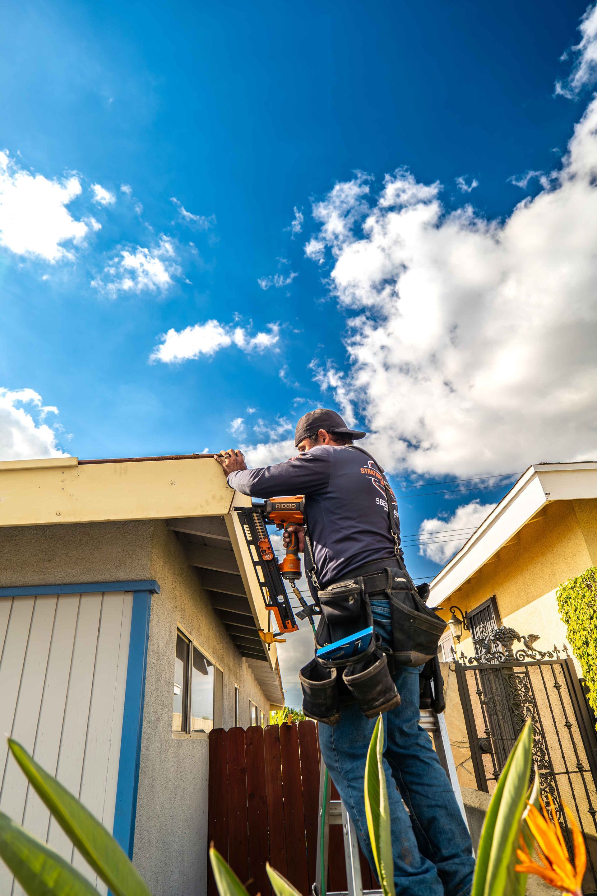 Roofer on ladder, working on a house roof under a blue sky with clouds.