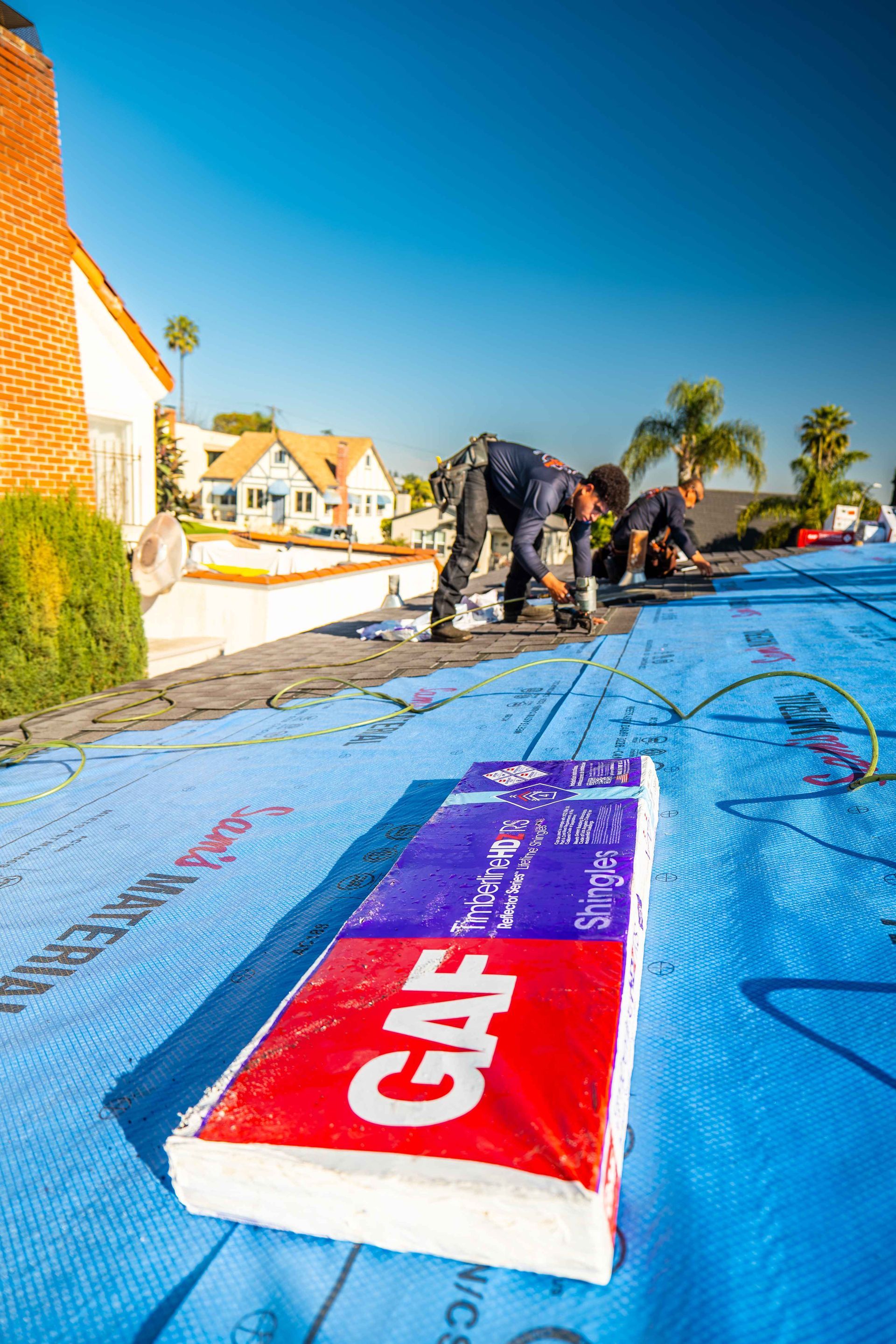 Roofers installing shingles on a house roof. GAF product box in foreground, blue tarp, sunny day.