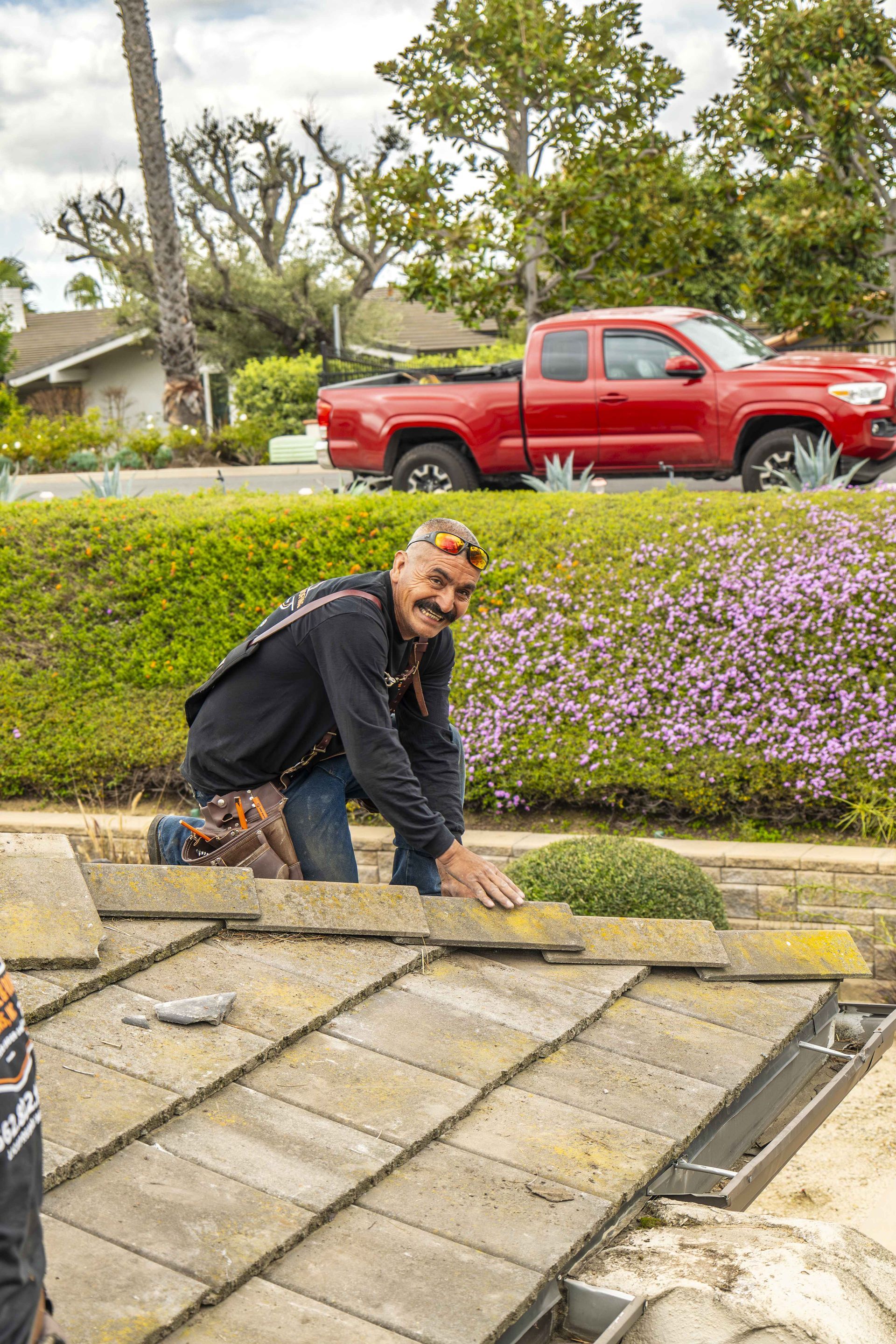 Man on a roof, installing tiles. Red truck and green shrubs in the background. Sunny day.