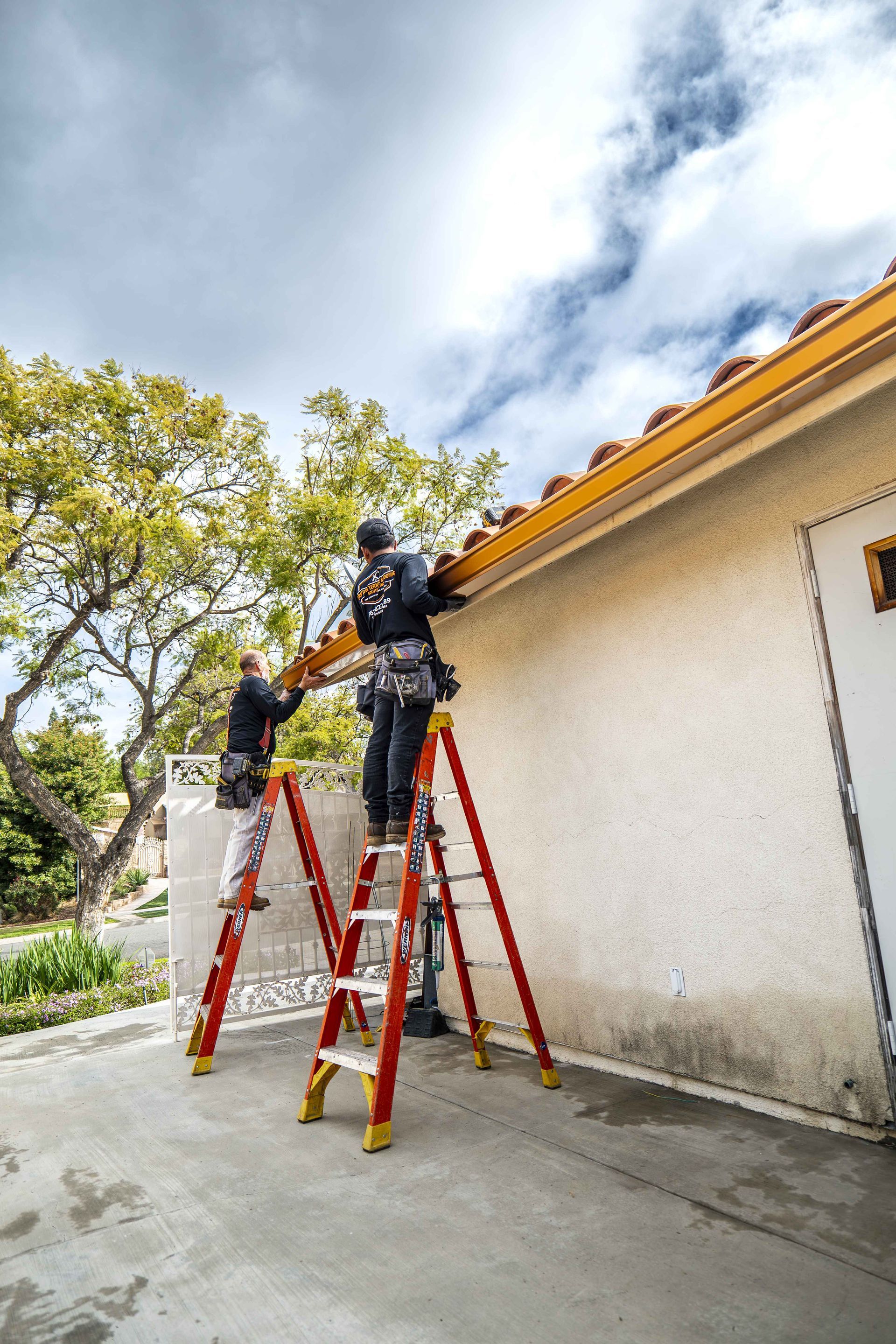 Two workers on ladders install gutters on a house under a cloudy sky.