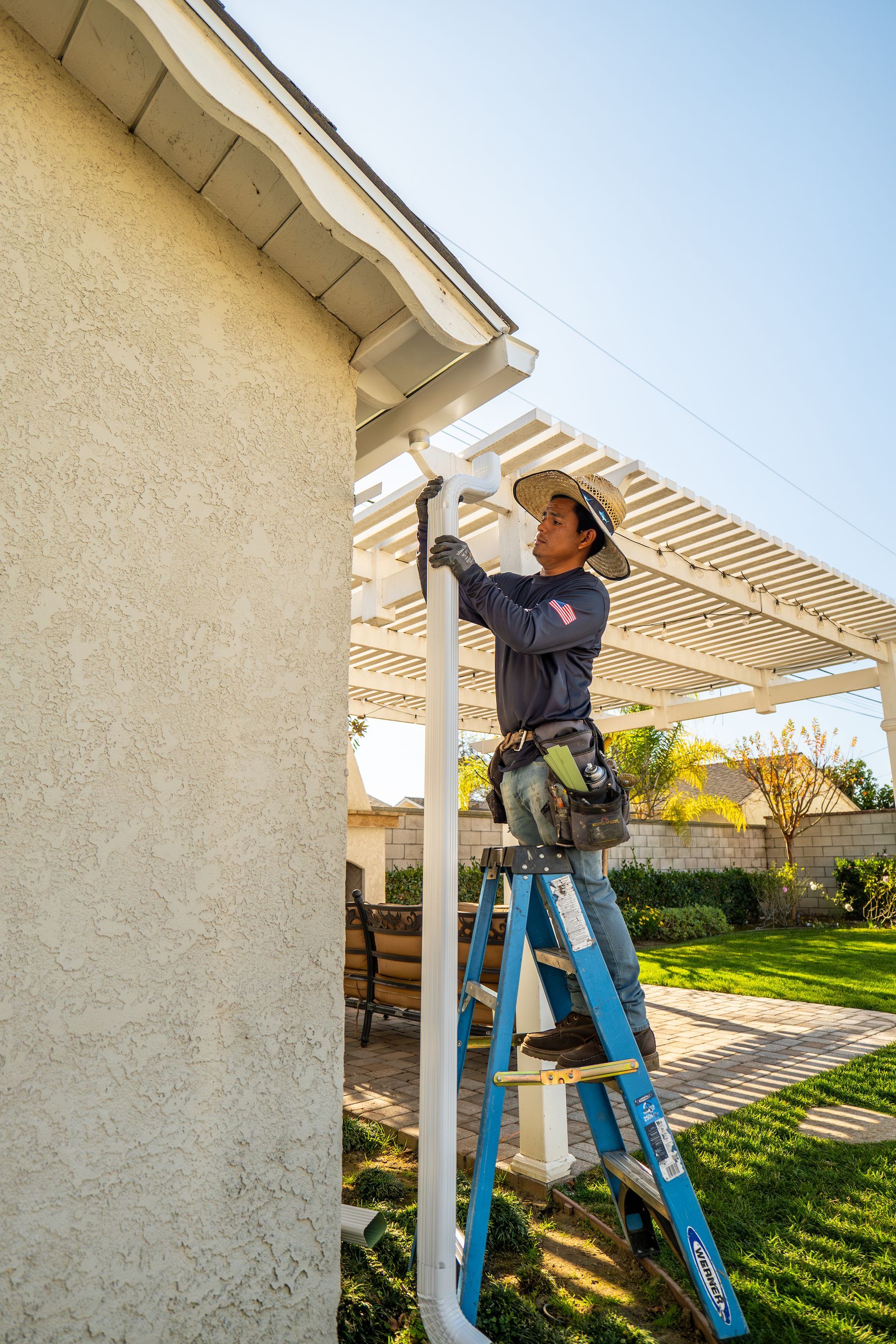 A man is standing on a ladder fixing a gutter on the side of a house.