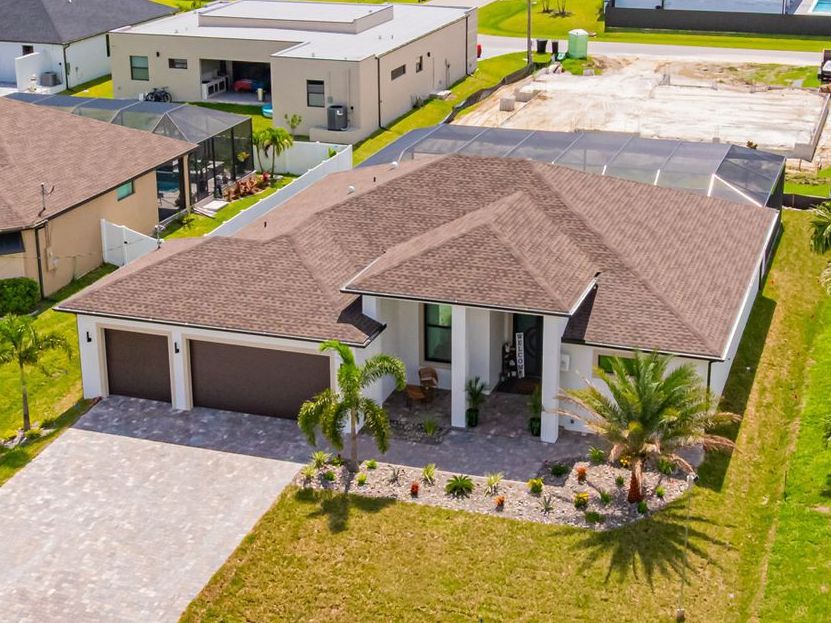 Aerial view of a modern, white house with a brown roof and a two-car garage, palm trees in front, and green lawn.