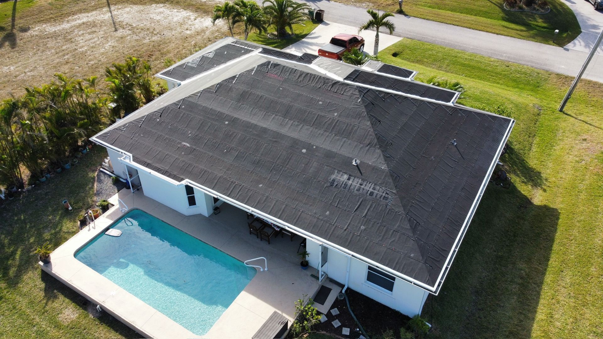 Aerial view of a white house with a black roof, pool, and green yard.