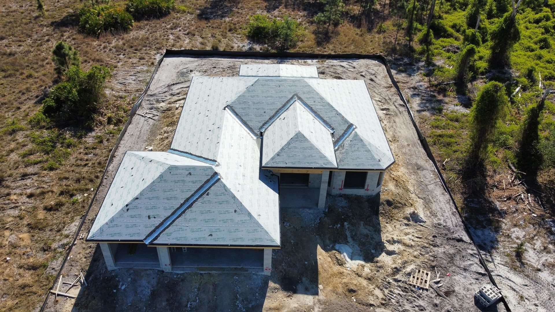 Aerial view of a house under construction with a gray roof and concrete foundation surrounded by dry land and sparse trees.