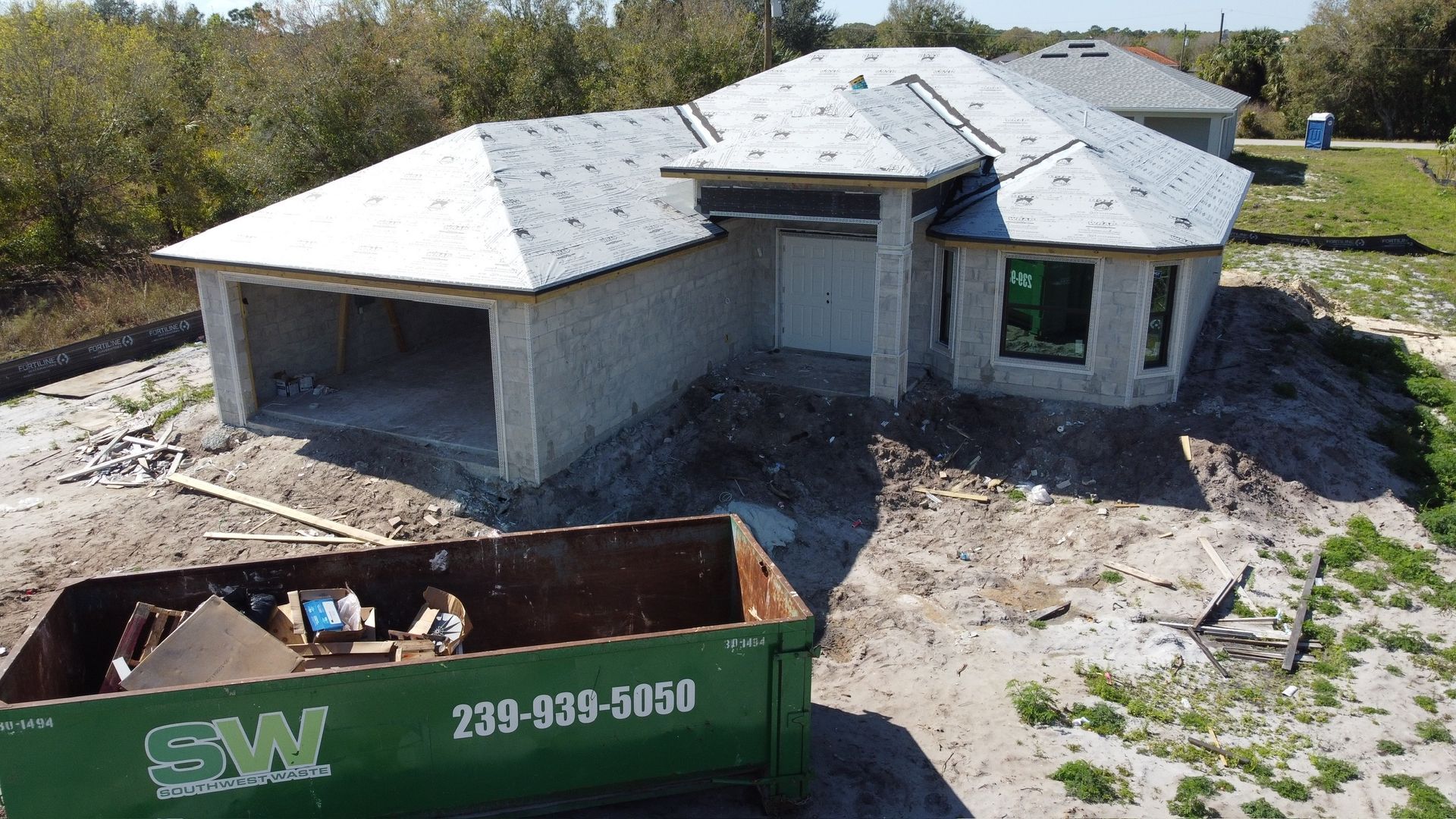 New home under construction; gray concrete block structure, green dumpster in foreground, sunny outdoor setting.