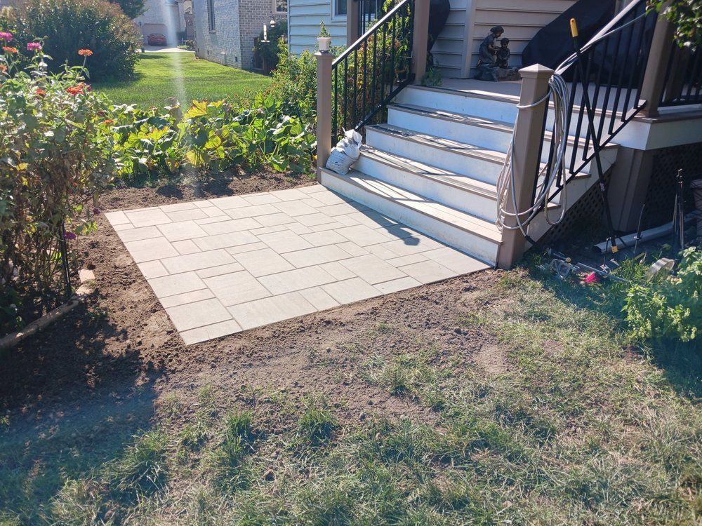 A square patio made of grey tiles sits at the base of wooden stairs, next to grass and garden beds.