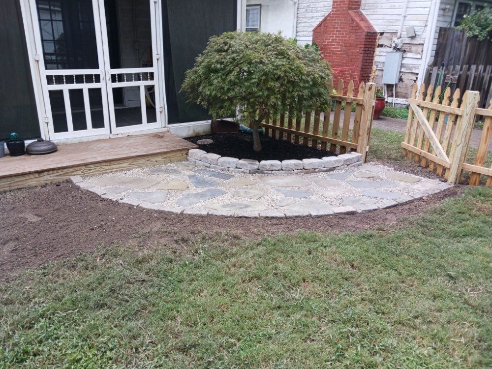 Stone patio with a small tree surrounded by a picket fence, in front of a house.