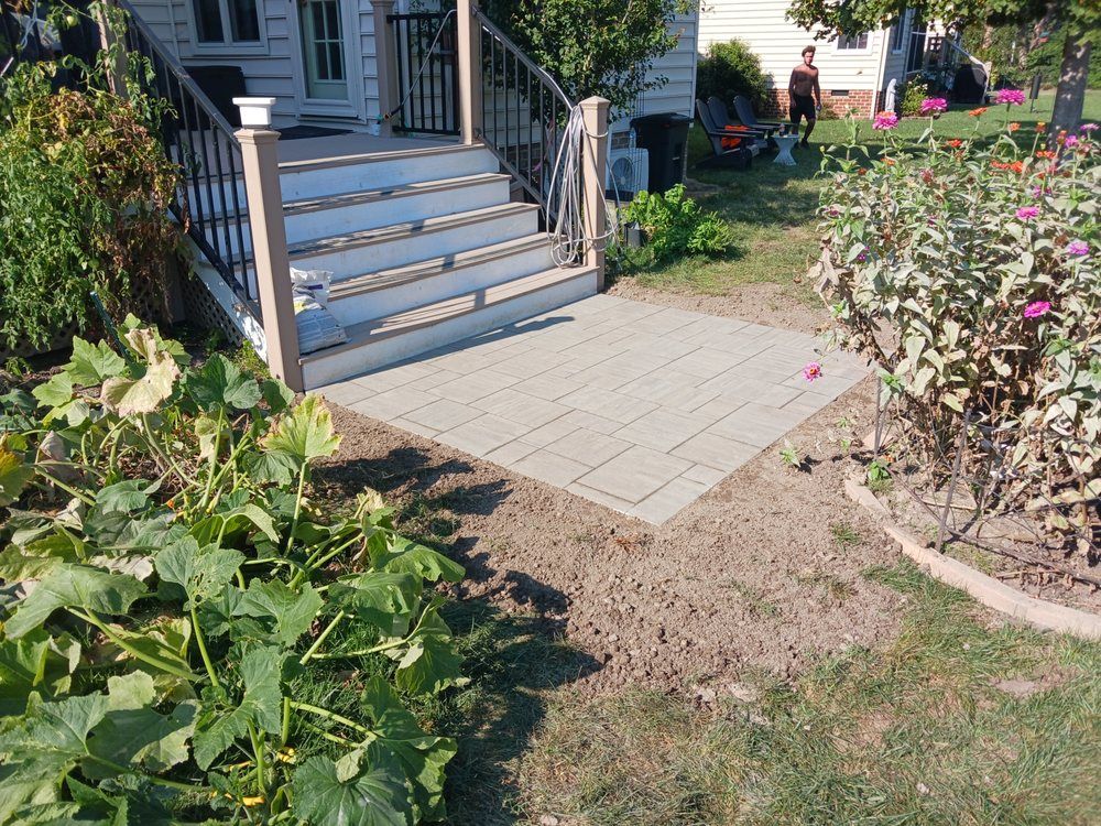 Stone patio in front of house steps with surrounding plants and person in the background.