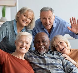 Five individuals smiling and looking at the camera, with one person waving, in a bright indoor setting.