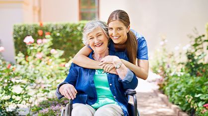 Smiling woman in a wheelchair with a young woman hugging her from behind in a garden path