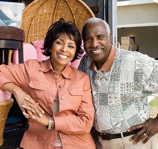 A smiling couple stands in front of a parked moving truck filled with furniture, including a large wicker chair.