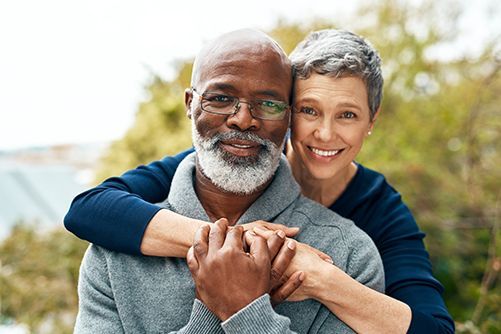 Smiling couple embracing outdoors, with trees and soft sunlight in the background