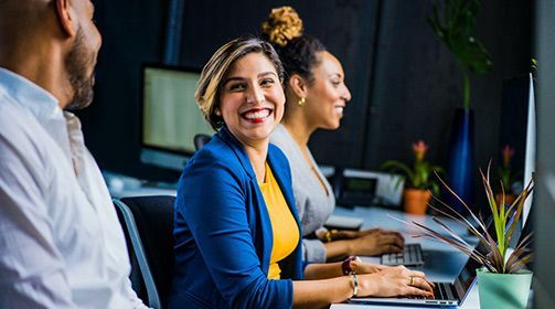 Three office workers collaborate at a desk, with a smiling professional in a blue blazer typing on a laptop.