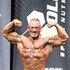 Bodybuilder flexing with arms up, showing defined muscles. Wearing no shirt, posing on stage.