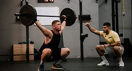 Man doing overhead squat with barbell, coach watching, gym setting.