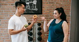 Man and woman in gym holding food. Man has a donut, woman points. Brick wall and weights in background.