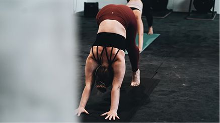 Woman in downward-facing dog yoga pose, wearing black top and burgundy leggings, in a studio.