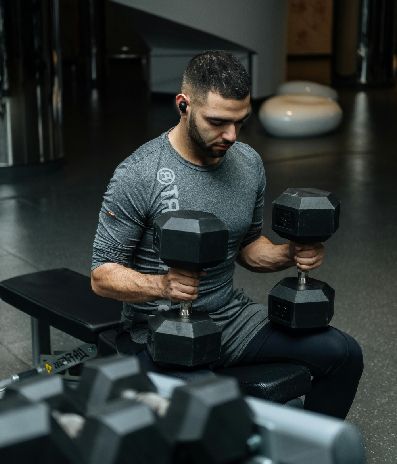 Man in gym, seated on bench, lifting dumbbells. Gray shirt, dark hair.