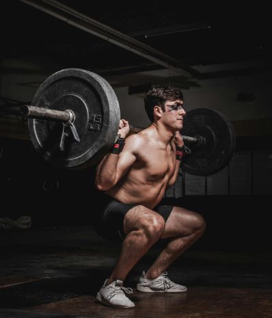Man squatting with a barbell in a dark gym. He's shirtless, wearing black shorts and white shoes.