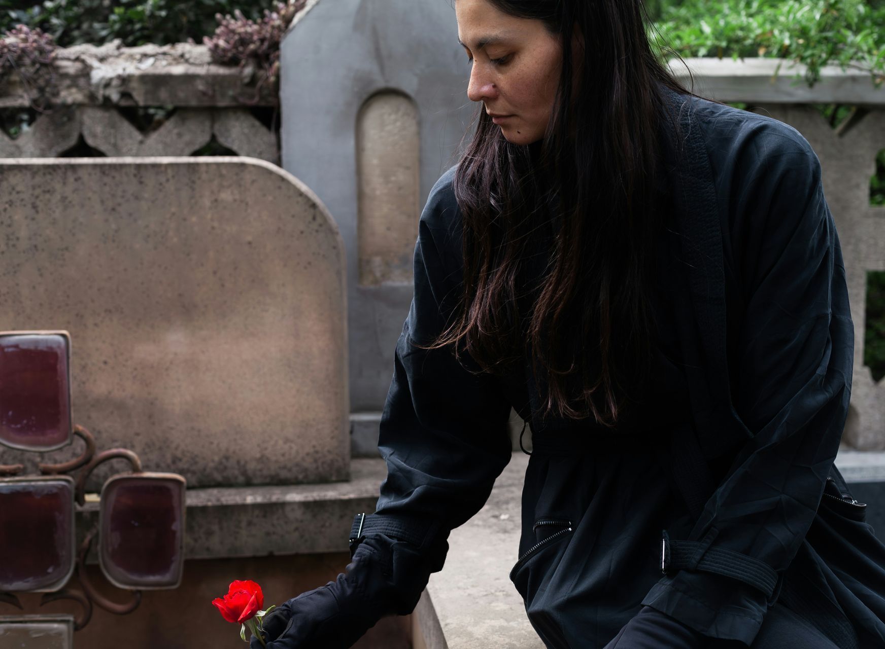 A woman is sitting in front of a grave holding a red rose.