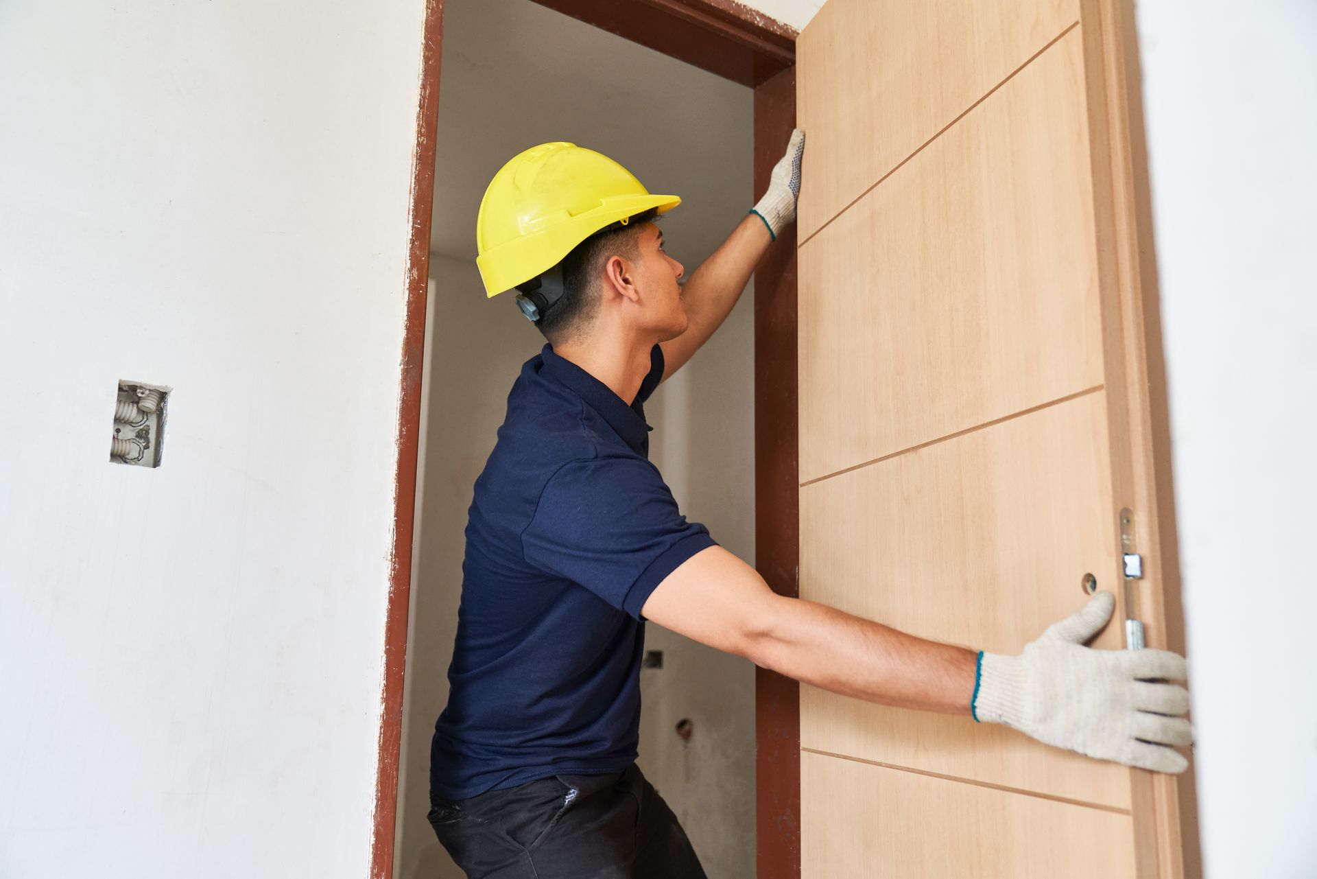 Worker in gloves and hard hat installing a wooden door in a doorway.