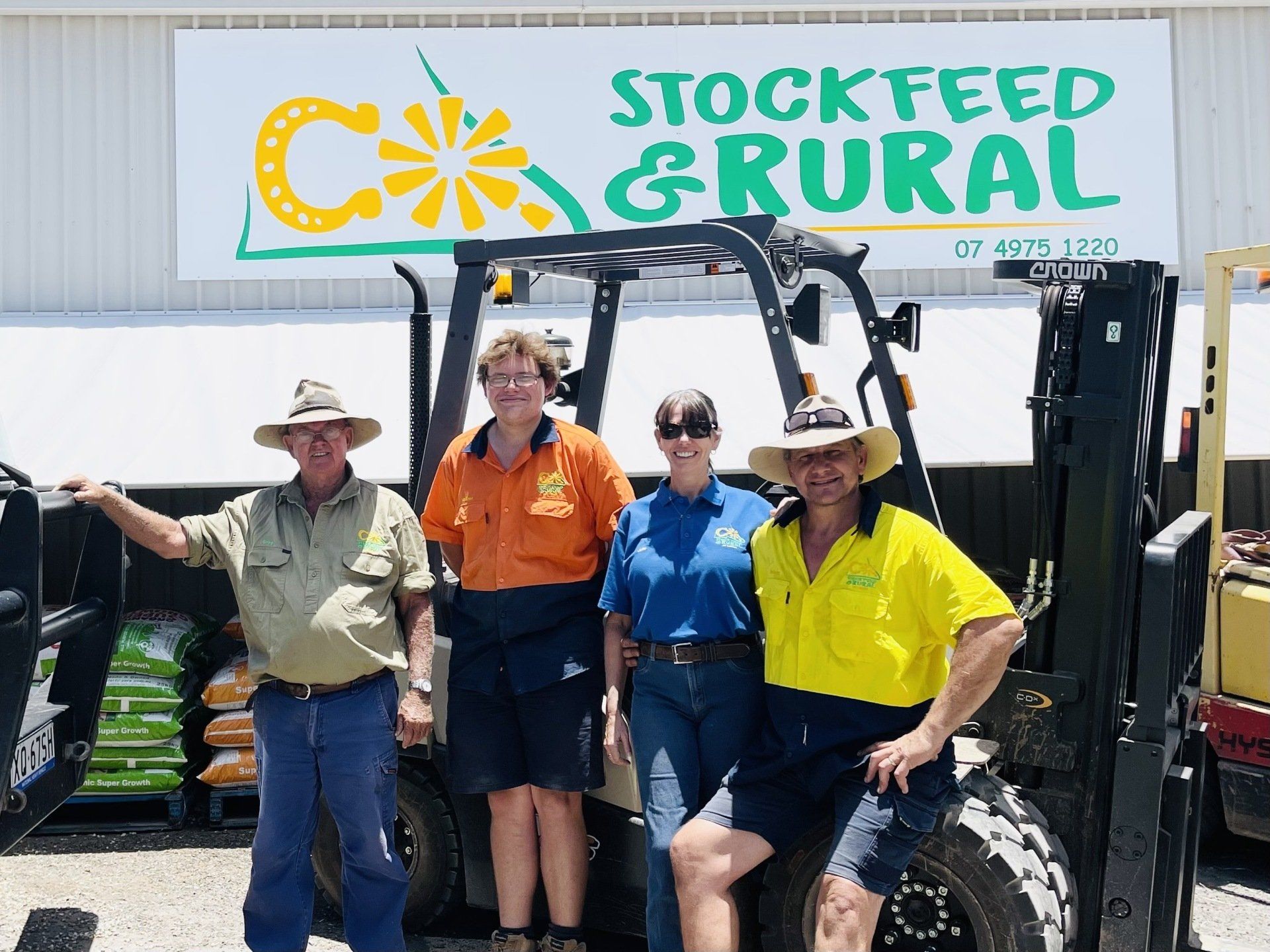 A group of people standing next to a forklift in front of a sign that says stockfeed & rural