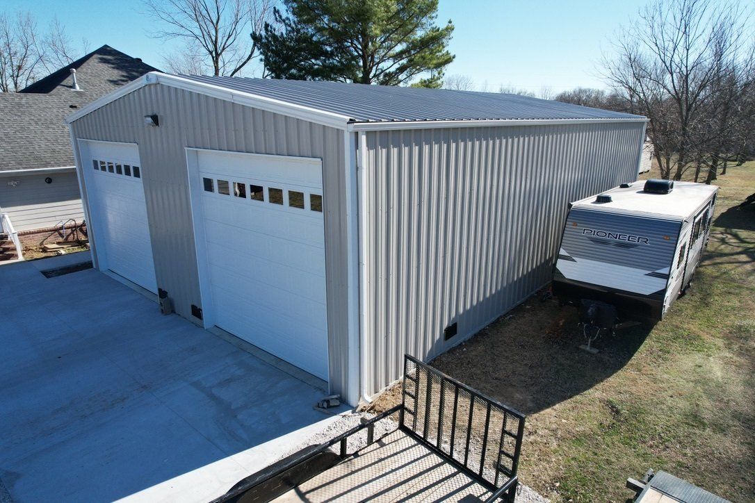 Gray metal building with two garage doors, an RV parked alongside it, and a small deck in the foreground.