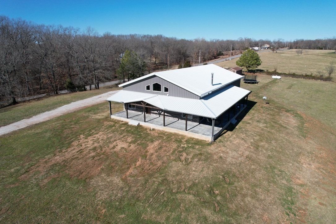 An aerial view of a gray, rectangular house with a white roof and covered porch in a rural setting. A gravel driveway leads up to the house.