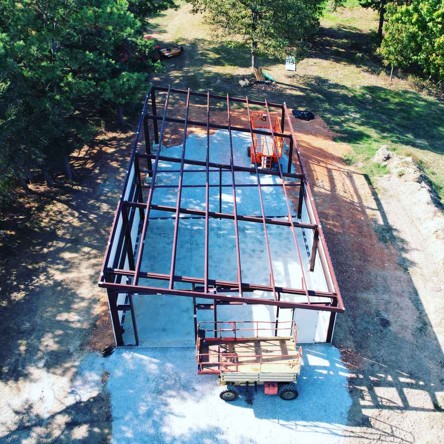 Aerial view of a metal frame building under construction. A lift is parked in front; the structure sits on a concrete pad surrounded by dirt and trees.