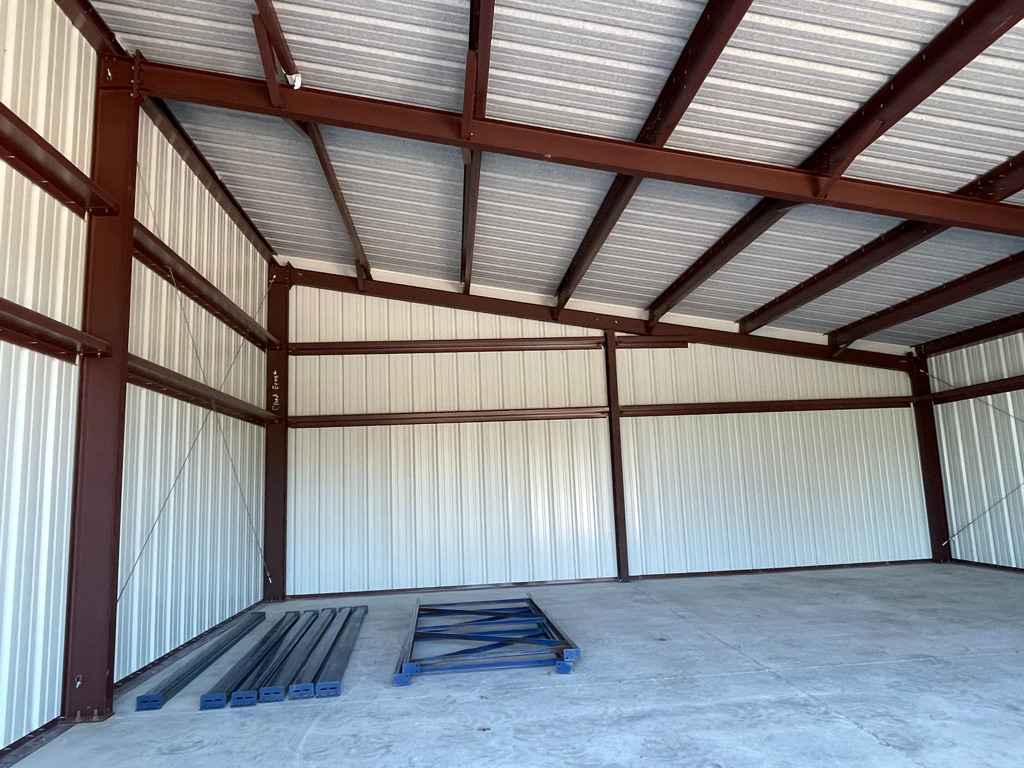 Interior of a brown metal frame building with a corrugated metal roof and walls, a concrete floor, and some disassembled metal pieces.