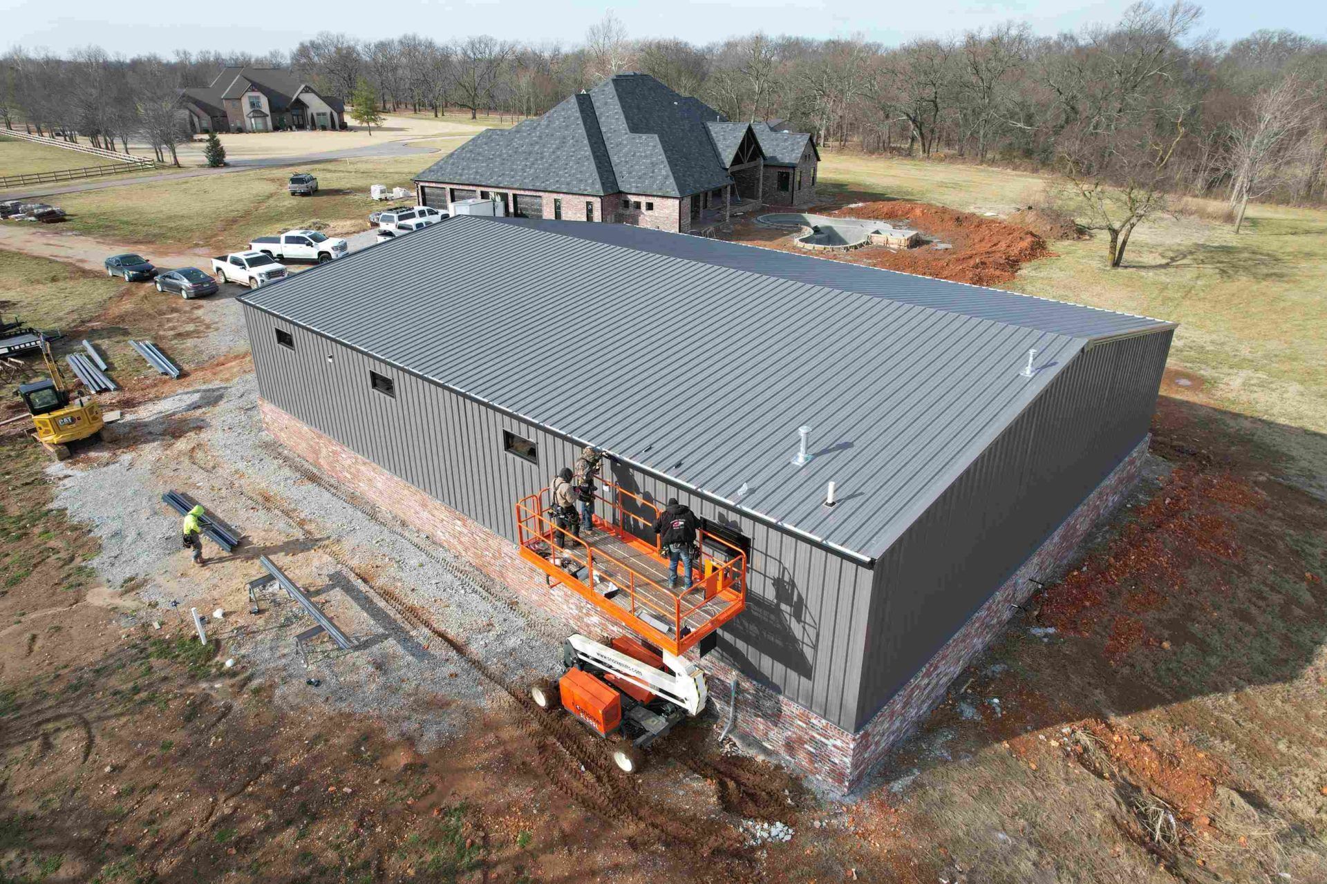 Construction of a large gray metal building with a brick base; a lift platform is in use. A house is in the background.