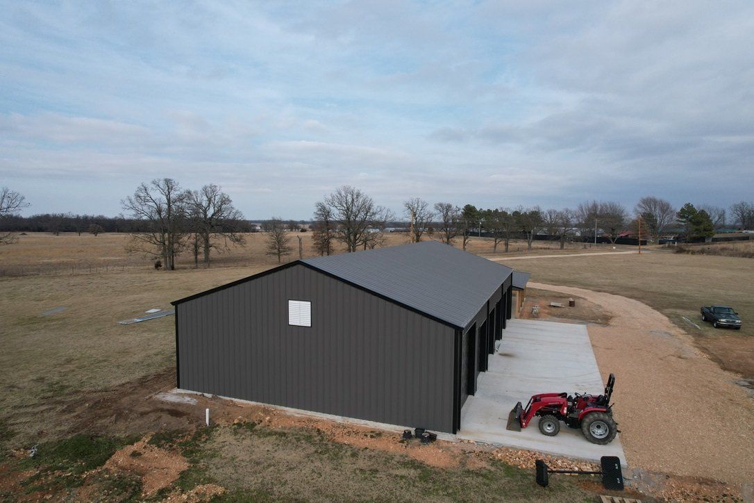 Dark gray metal building with multiple garage doors, concrete pad, and a tractor on a gravel drive in a rural setting under a cloudy sky.