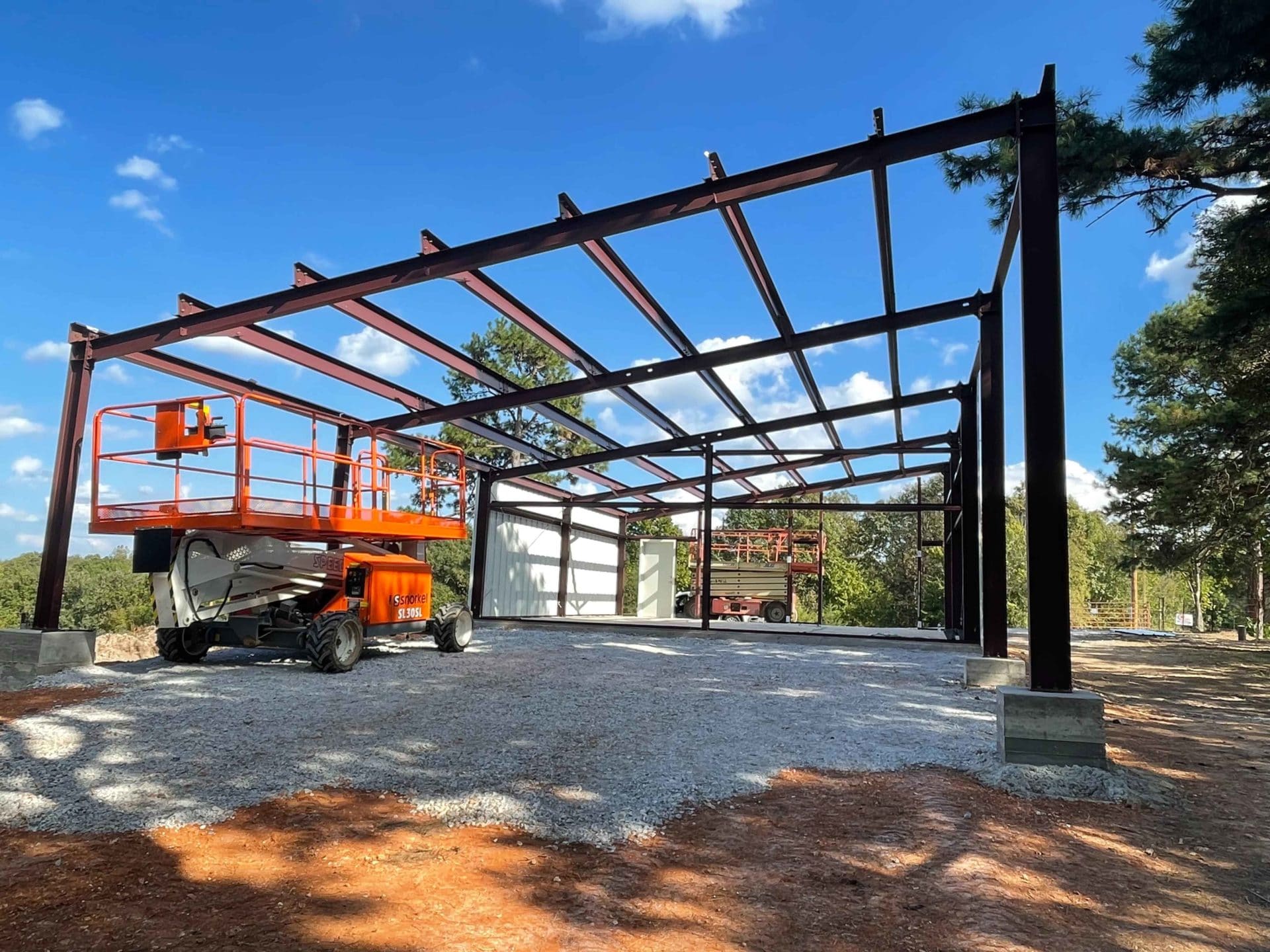 Steel frame of a building under construction with a cherry picker, on a gravel foundation, against a backdrop of trees and a blue sky.